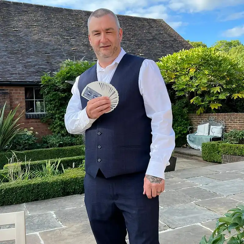 Main character of image is Tony Byrne, tony is wearing smart dress, holding playing cards in a fan. Tony is outside on a patio.